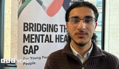 Rushid has long black hair and a beard and is wearing glasses, a blue and white shirt and a brown zipped top. He is standing in front of a white banner which says "bridging the mental health gap" in black letters.