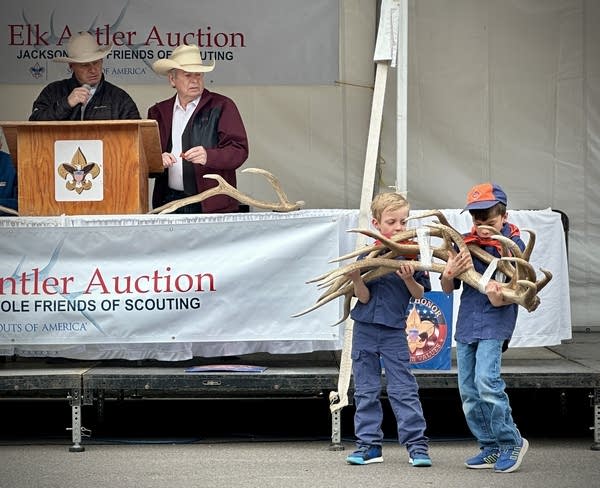 Two boys carry a bundle of antlers while two auctioneer men in cowboy hats look on.