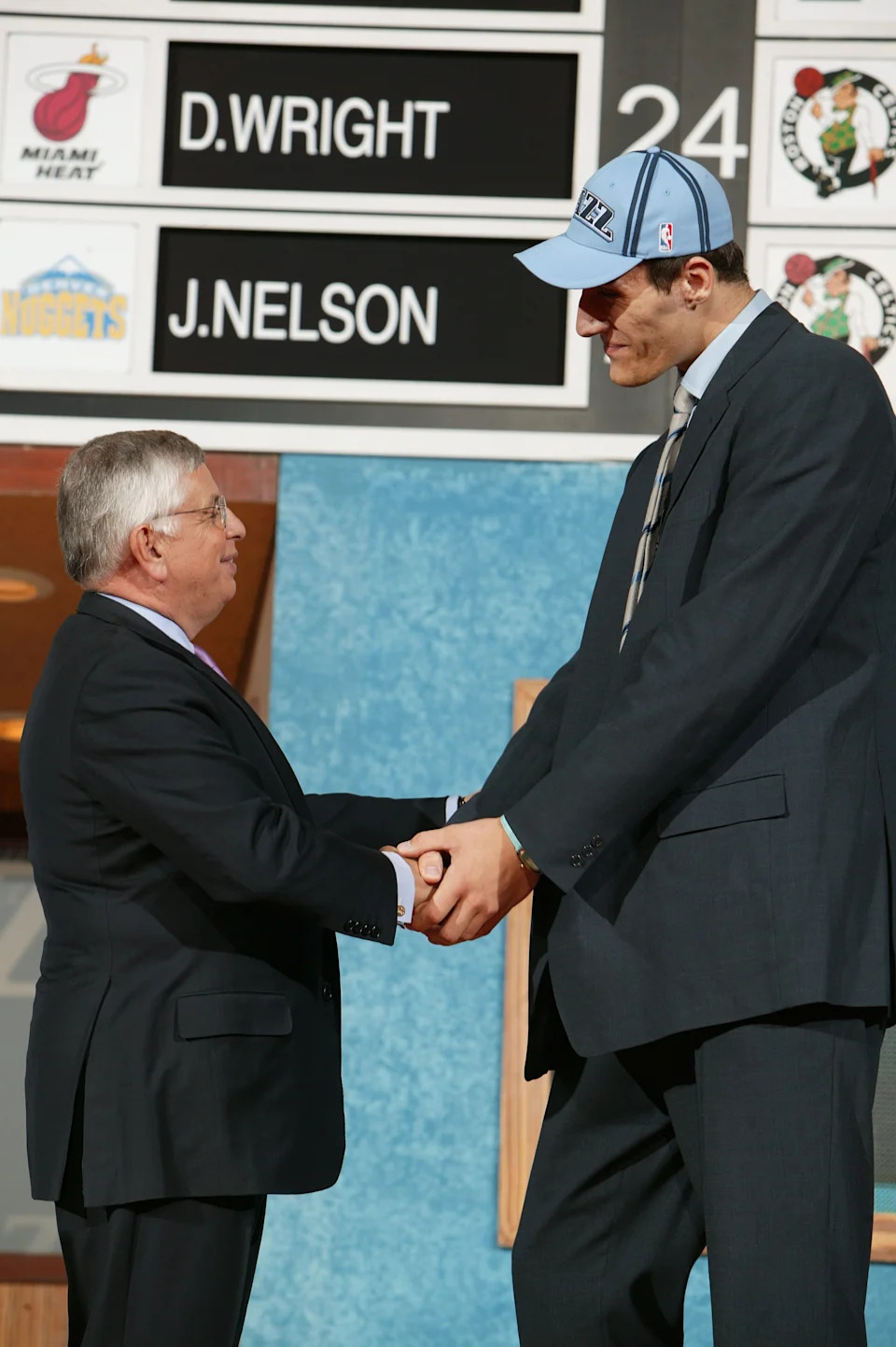 A tall basketball player in a suit is shaking hands with an official on a stage under a draft board during NBA Draft proceedings