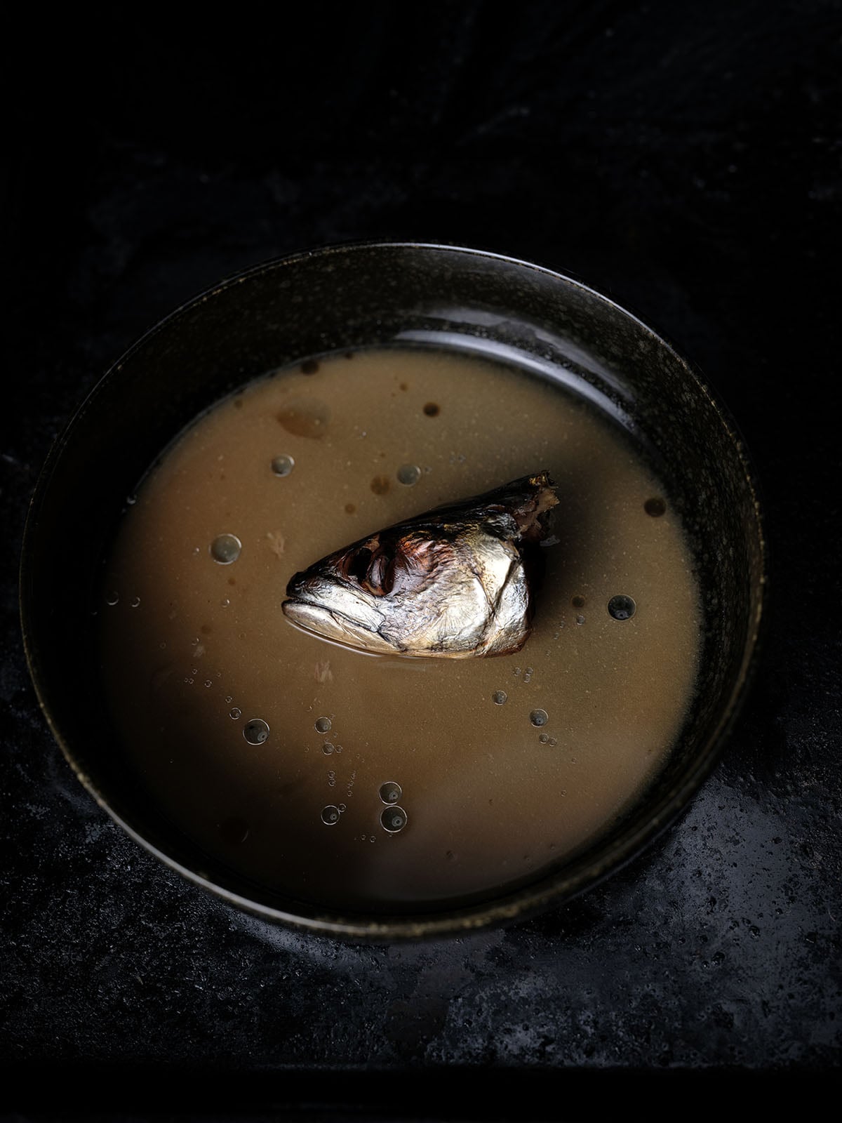 A dark bowl filled with brown soup, with a cooked fish head floating in the center, sits on a dark, textured surface.