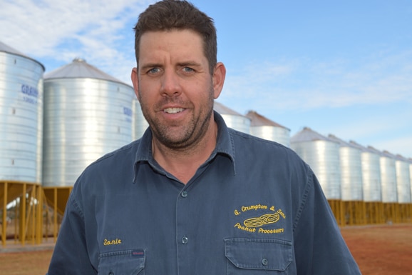 A man in a blue work shirt stands in front of silver silos against a blue sky.