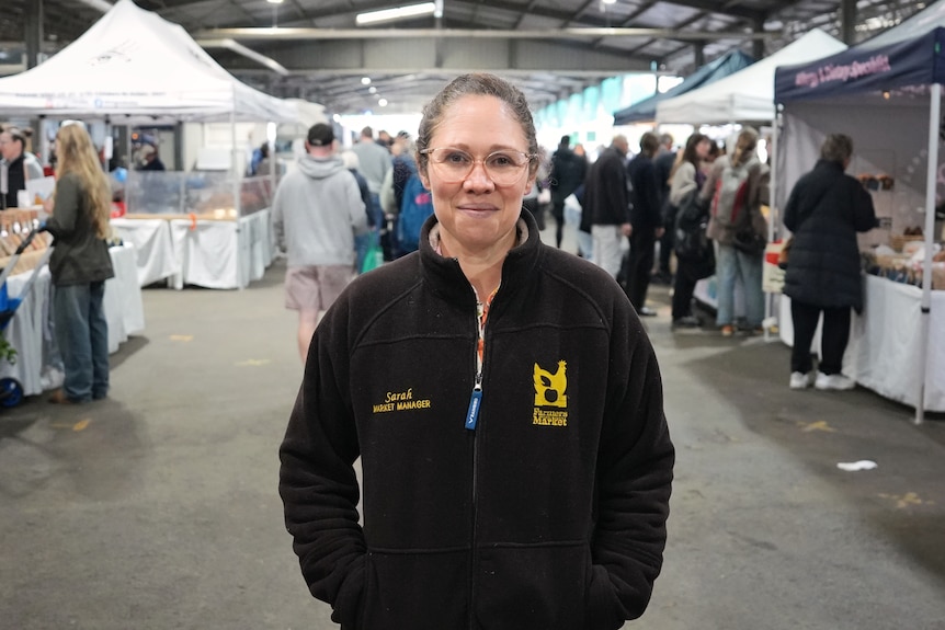 A woman with light hair pulled into a ponytail stands in a warehouse surrounded by market stalls.