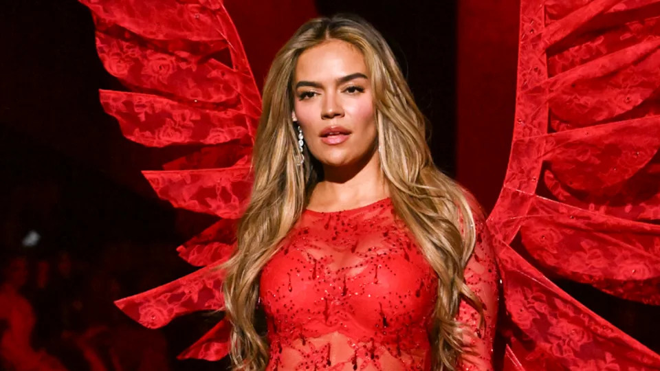 Karol G with long hair in a red lace outfit and matching wings stands confidently on stage.