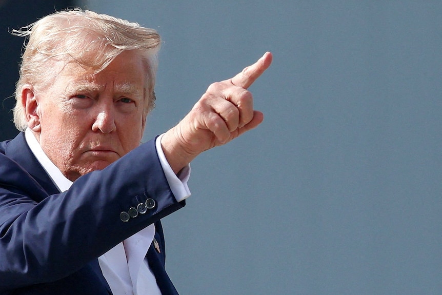 A man in a dark suit — Donald Trump — points to the sky.