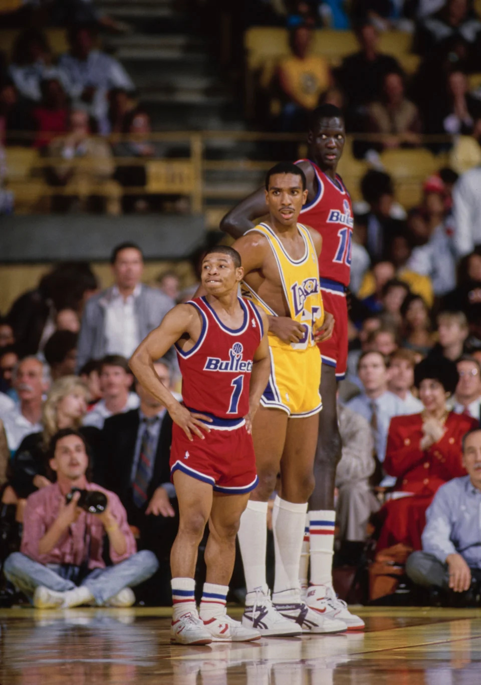 Basketball players on a court, with one player notably shorter. Spectators fill the stands in the background