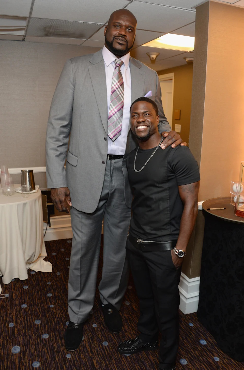 Two men are posing together, one very tall in a suit and the other shorter in a casual outfit, smiling indoors