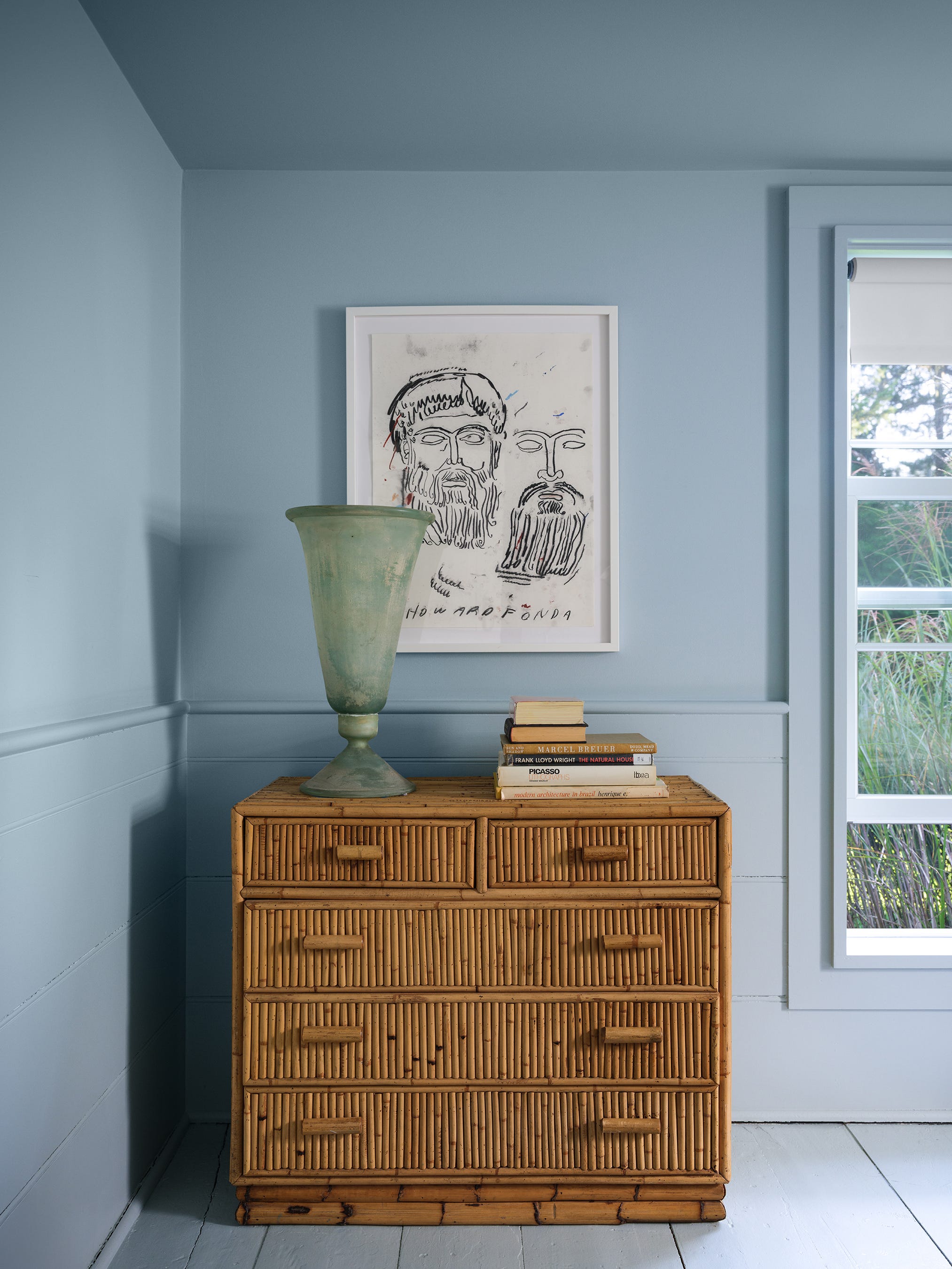 Rattan dresser with books and a vase, blue wall, abstract art, and window view.