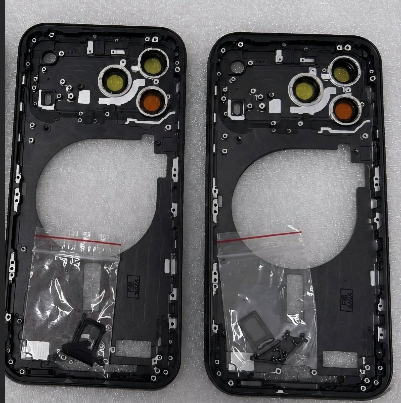 Two identical black smartphone back frames with three circular camera cutouts each, lying side by side on foam, with small clear plastic bags containing tiny components near the bottom.