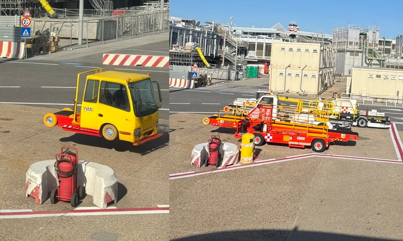 Airport service vehicles on a tarmac, including small yellow and red tow trucks and equipment carts, with safety barriers, gas cylinders, and industrial buildings in the sunny background