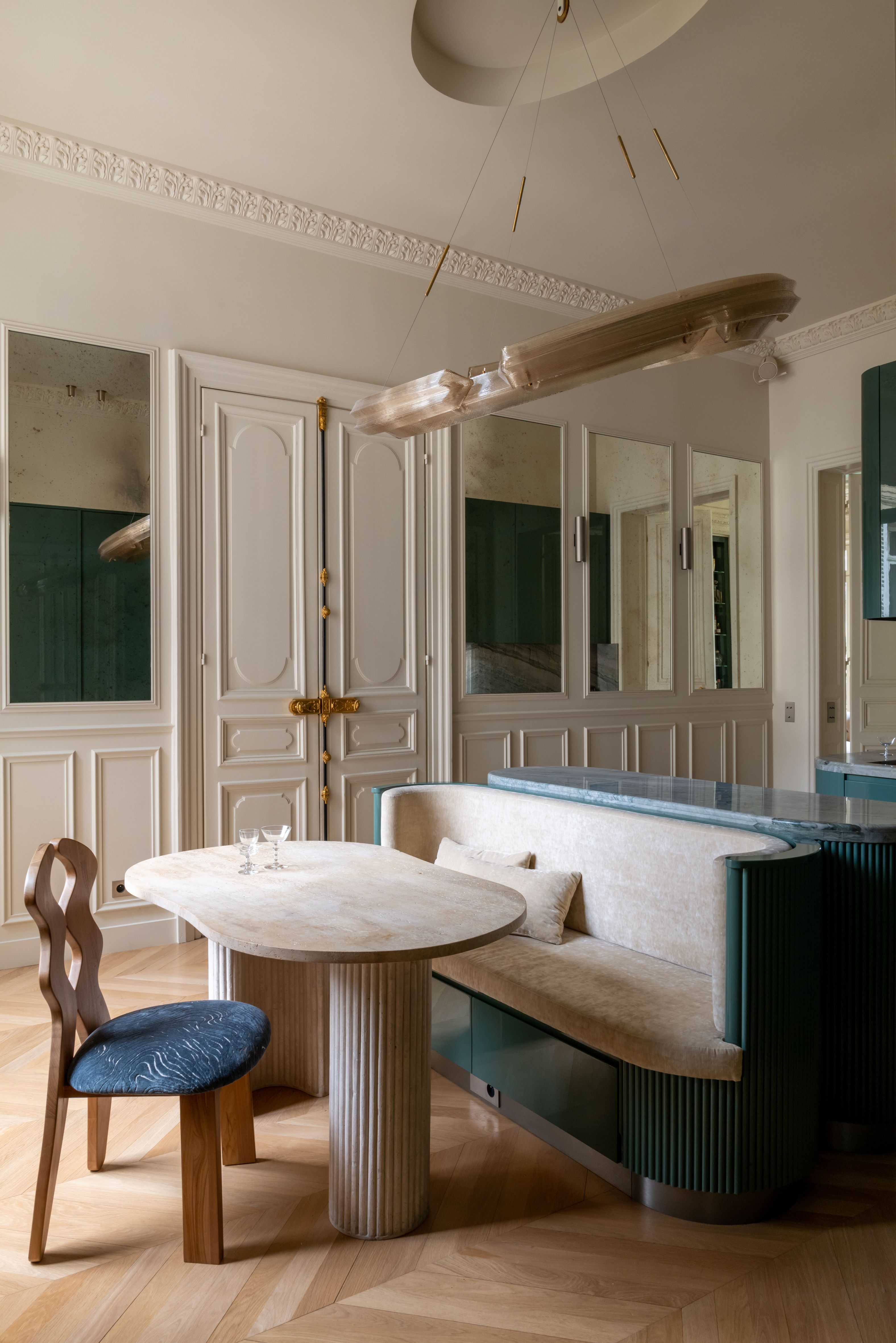 A grand dining room with off white walls, architectural moldings and paneling, including mirror panels, with an off-center circular pendant light above a banquette and table