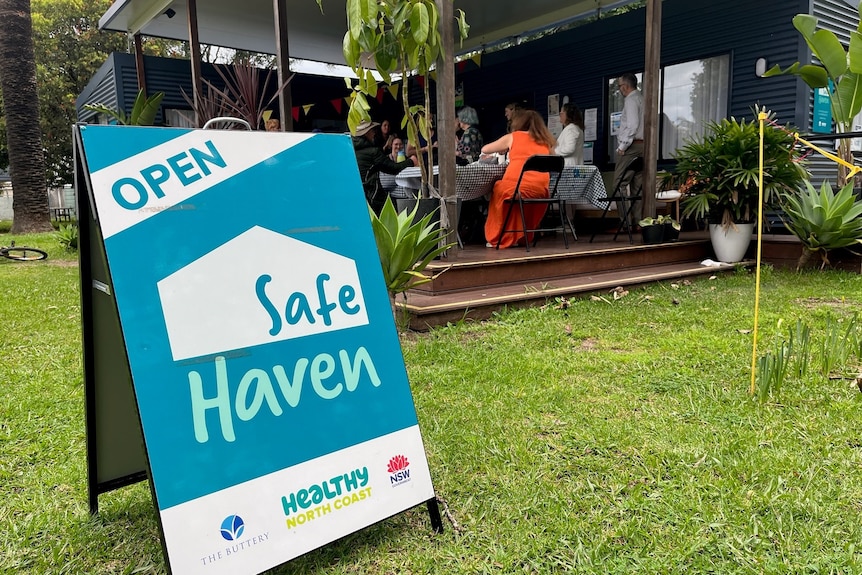 Healthy North Coast sign on grass which says open safe haven, a lady in orange behind sign sits at table of pop-up clinic.