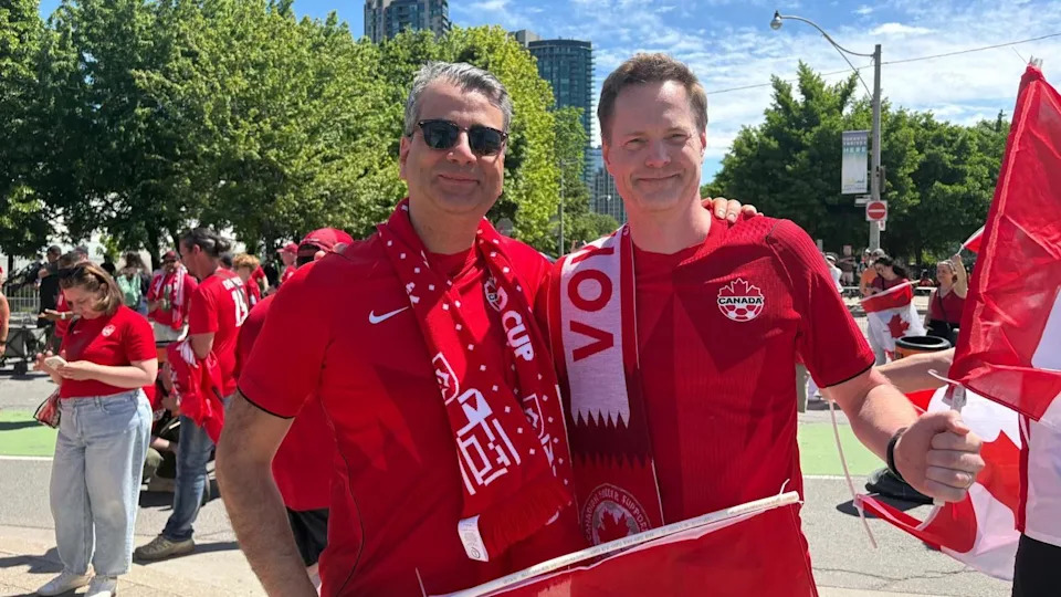 An image of two men side-by-side wearing Team Canada jerseys and Team Canada scarves, waving Canadian flags. Behind them are other fans in wearing red and holding Canadian flags.