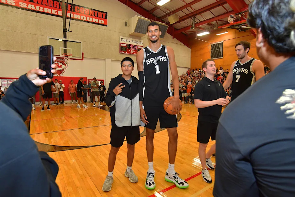 Basketball player in Spurs jersey poses with a fan on a gym court, surrounded by team and crowd