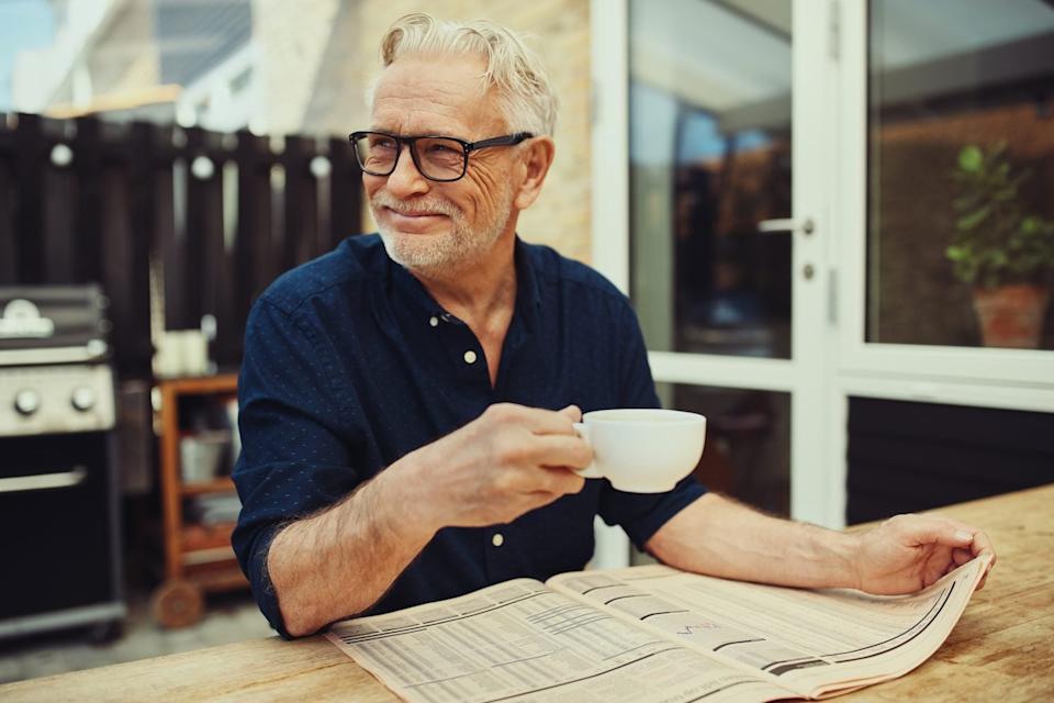 A smiling older man with grey hair, a beard, and glasses sits at an outdoor wooden table, holding a white coffee mug in his right hand and reading a newspaper spread open on the table. He is wearing a dark blue collared shirt. A black grill is visible in the blurred background on the left, and a white door with a window showing greenery is on the right.