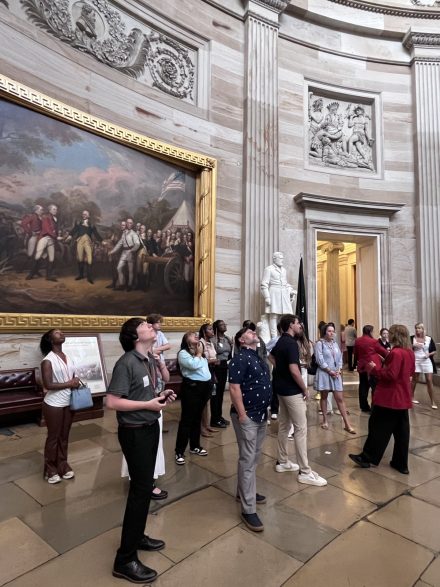 A group of students and staff stand inside the U.S. Capitol Rotunda, listening to a guide while looking up at the building’s artwork and architecture. A large historical painting and ornate marble walls are visible in the background.