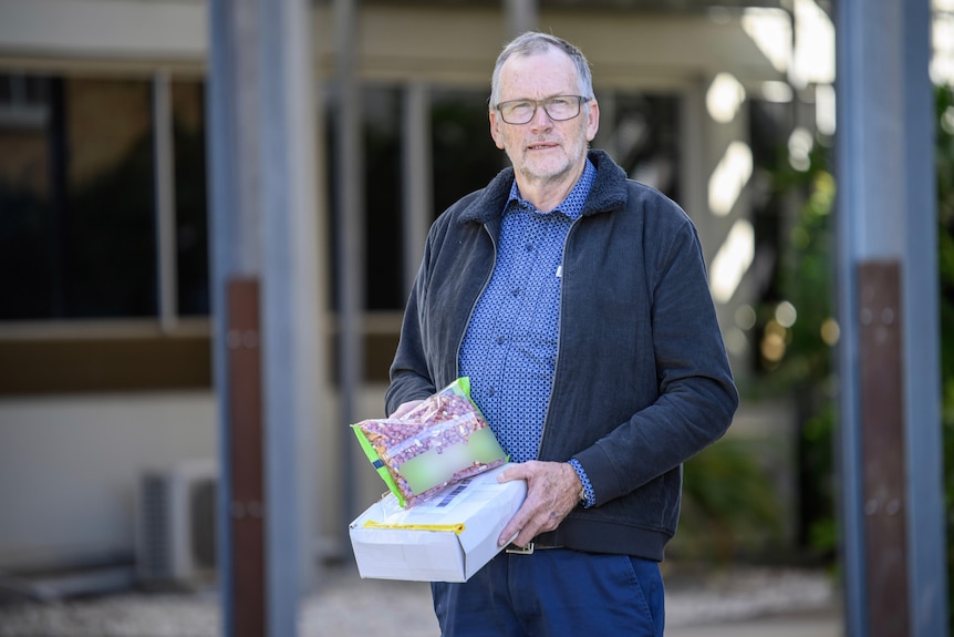 Graeme, with grey hair and a serious expression, holds a packet of peanuts and a box.