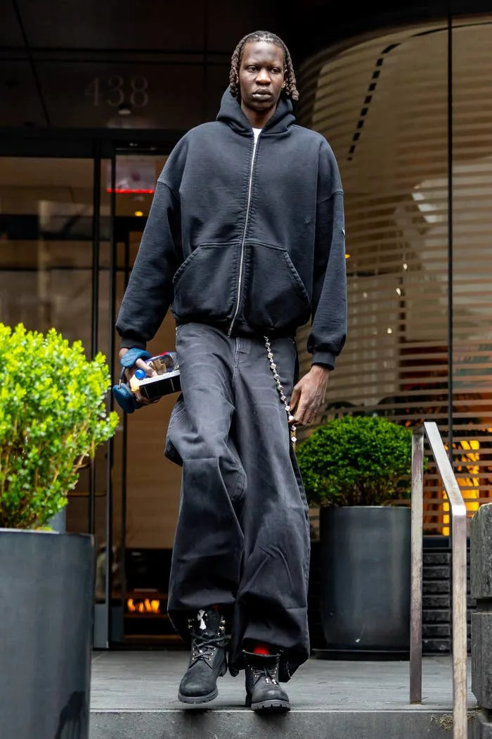 Person in oversized black hoodie and wide-leg pants, holding a water bottle, stepping outside a building, with plants nearby