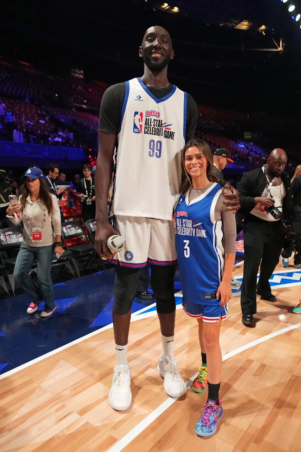 Two athletes in basketball jerseys at an All-Star Celebrity Game pose on a court, surrounded by fellow participants and media