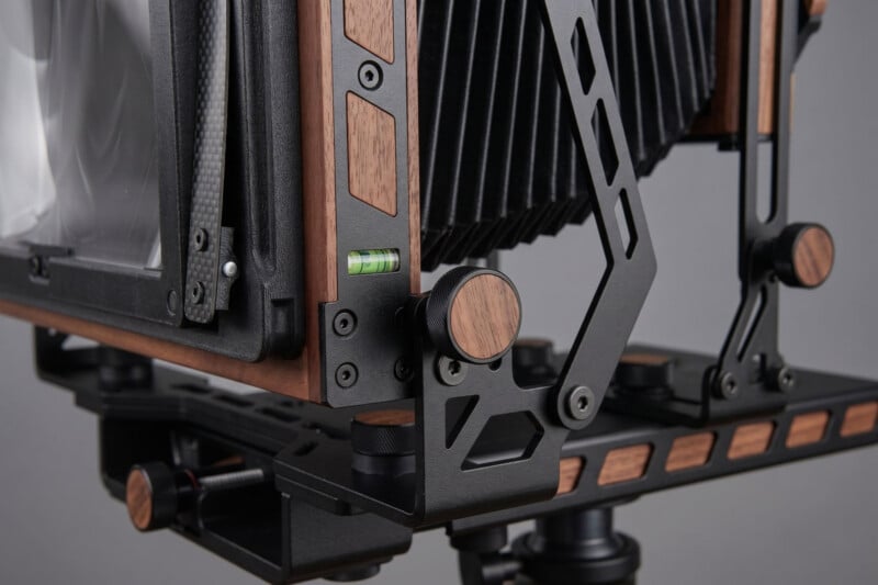 Close-up of a large format camera with a wooden and metal frame, bellows, adjustment knobs, and a small built-in spirit level, all mounted on a tripod.
