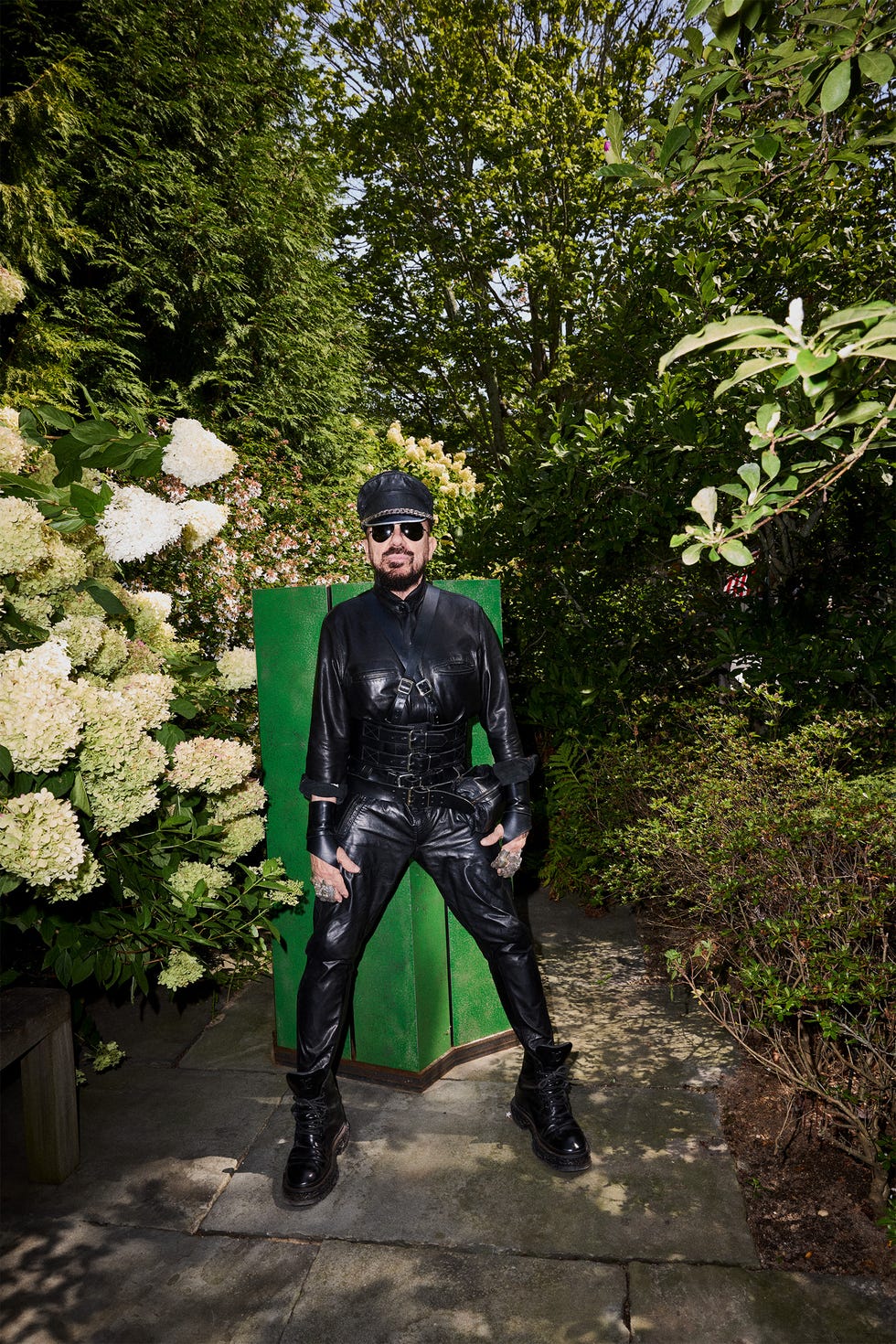Person in black leather outfit and sunglasses standing in a garden with green foliage.
