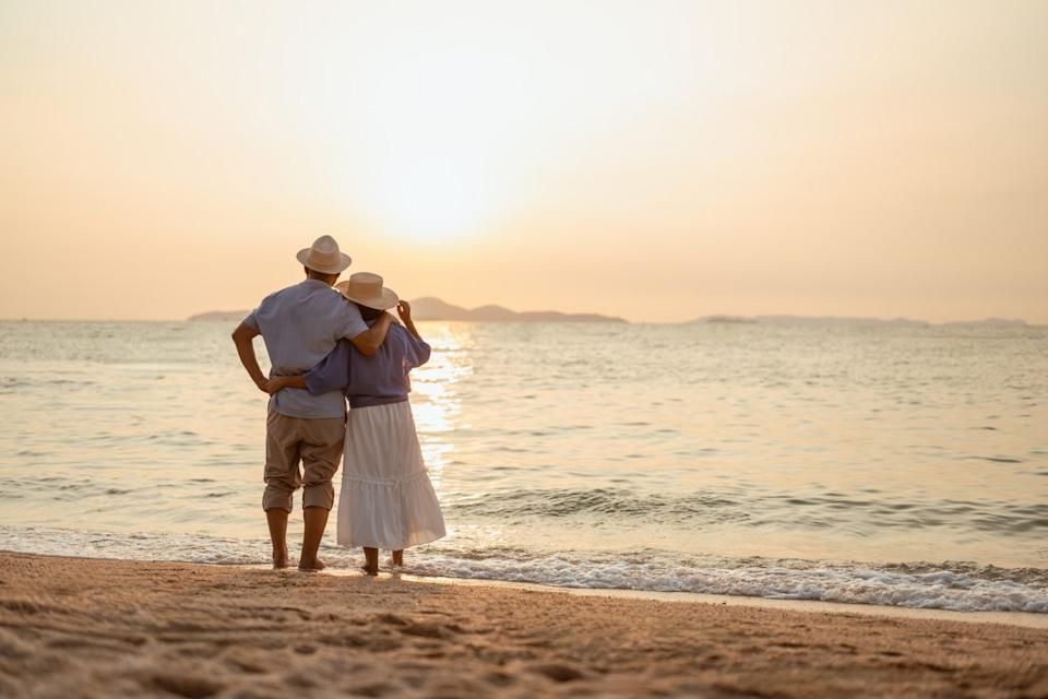 retired, retirement, couple, senior, elderly, beach, recreation, insurance, planning, vacation. A couple standing on the beach at sunset, with the man holding the woman's hand.