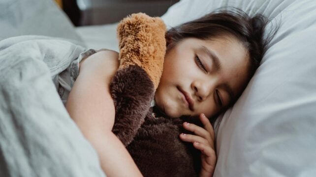 A young girl peacefully sleeping in bed with a stuffed teddy bear, showcasing tranquility and comfort.