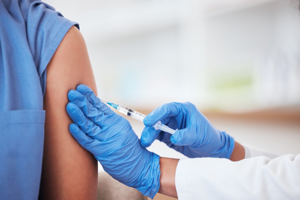 A person in blue scrubs receiving a vaccine injection in their arm from a gloved hand holding a syringe.