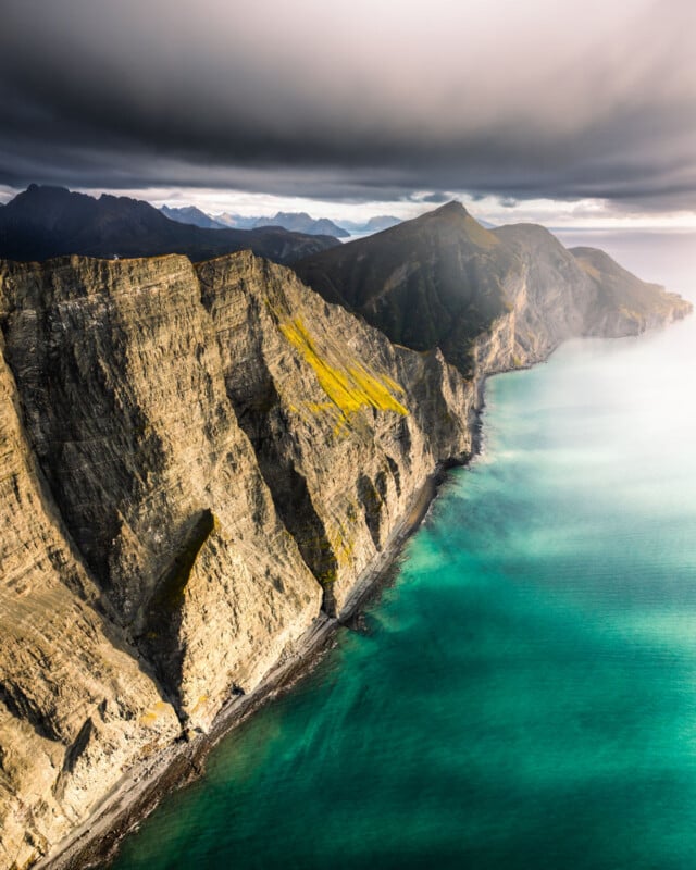 Dramatic cliffs rise sharply from turquoise sea water under a moody, cloudy sky, with mountains visible in the background and sunlight illuminating parts of the rocky landscape.