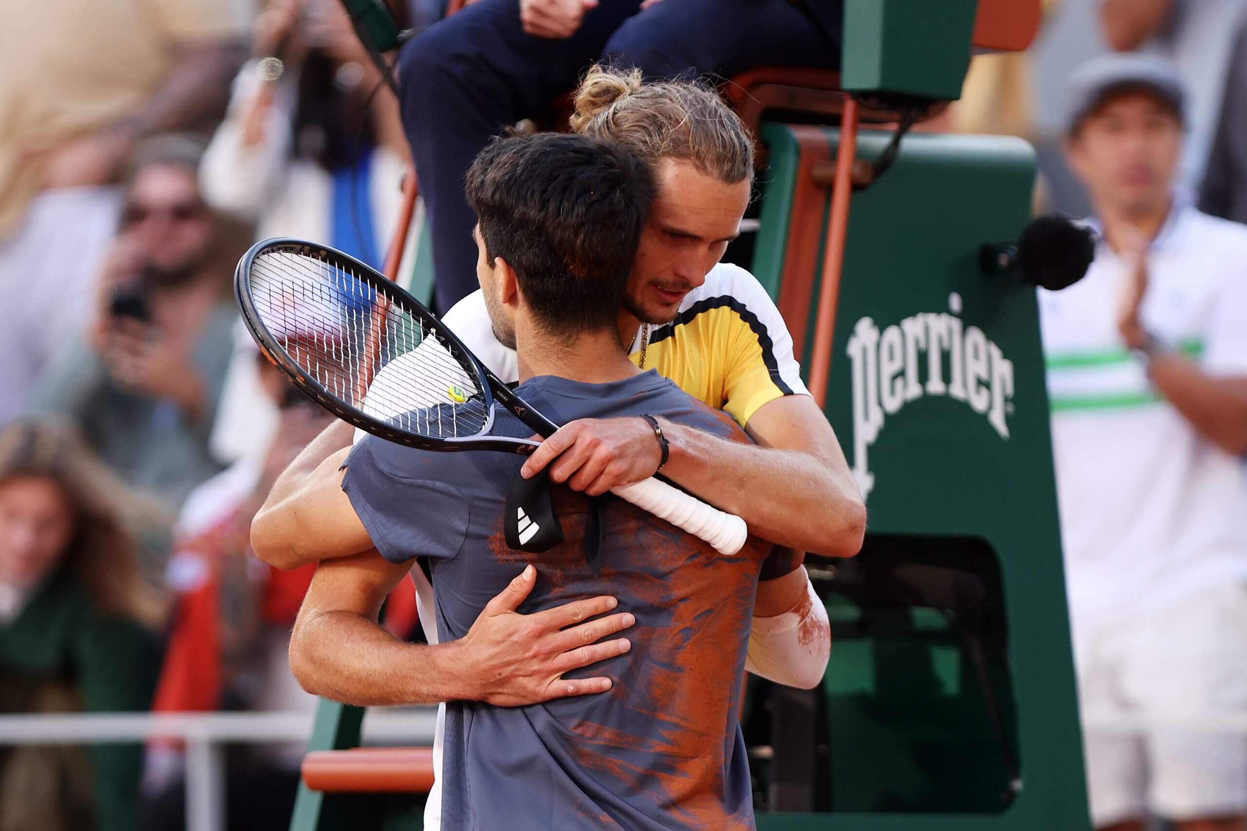 Alexander Zverev hugs Carlos Alcaraz at the end of a tennis match. Zverev wears a yellow Adidas kit as Alcaaz's back is streaked with clay.