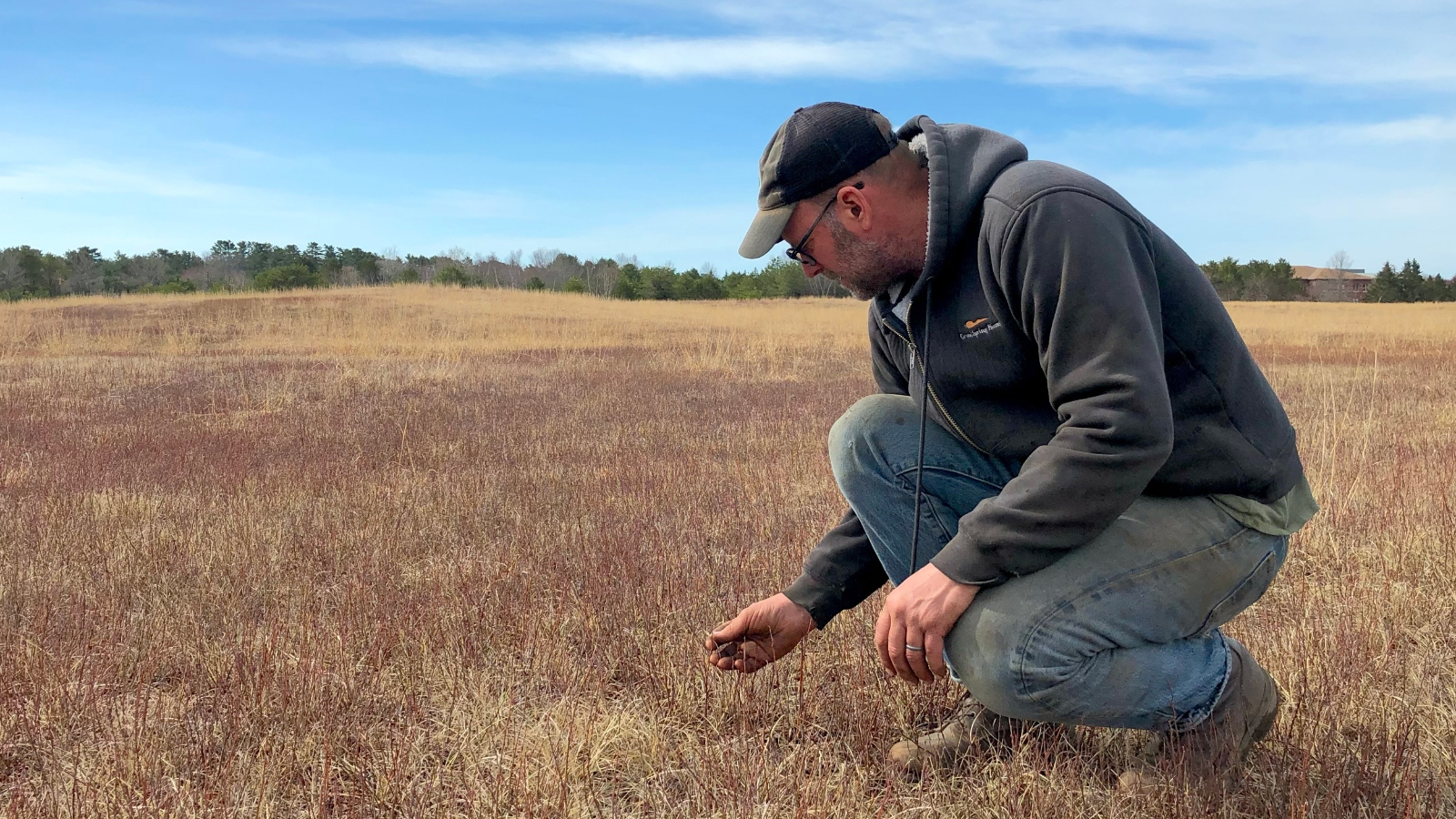 A man in jeans, a hoodie and baseball cap kneels in a parched field