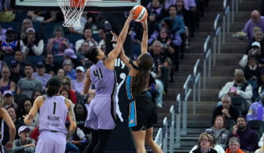 Two female basketball players jump near the hoop, reaching for the ball, while an audience watches the WNBA game from the stands.