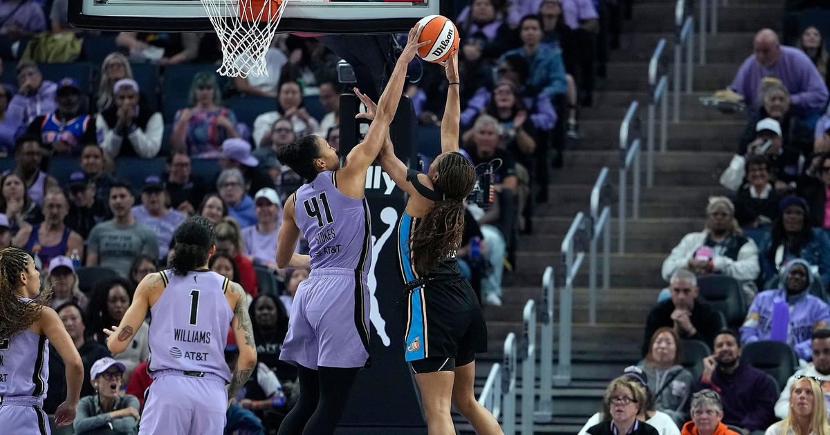 Two female basketball players jump near the hoop, reaching for the ball, while an audience watches the WNBA game from the stands.