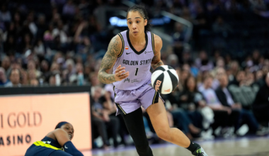 A basketball player in a purple Golden State uniform dribbles the ball swiftly past a fallen opponent in a dark blue uniform on the court.