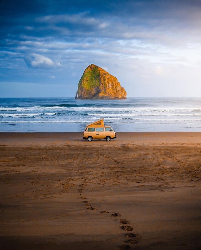 A tan camper van is parked on a sandy beach with footprints leading to it. In the distance, a large rocky island rises from the ocean under a partly cloudy sky with sunlight illuminating the rock.