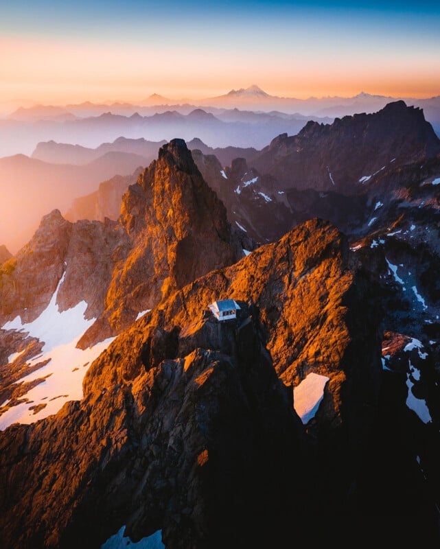 A small mountain cabin sits atop a rugged, rocky peak with patches of snow, surrounded by dramatic mountain ranges under a colorful sunrise or sunset sky.