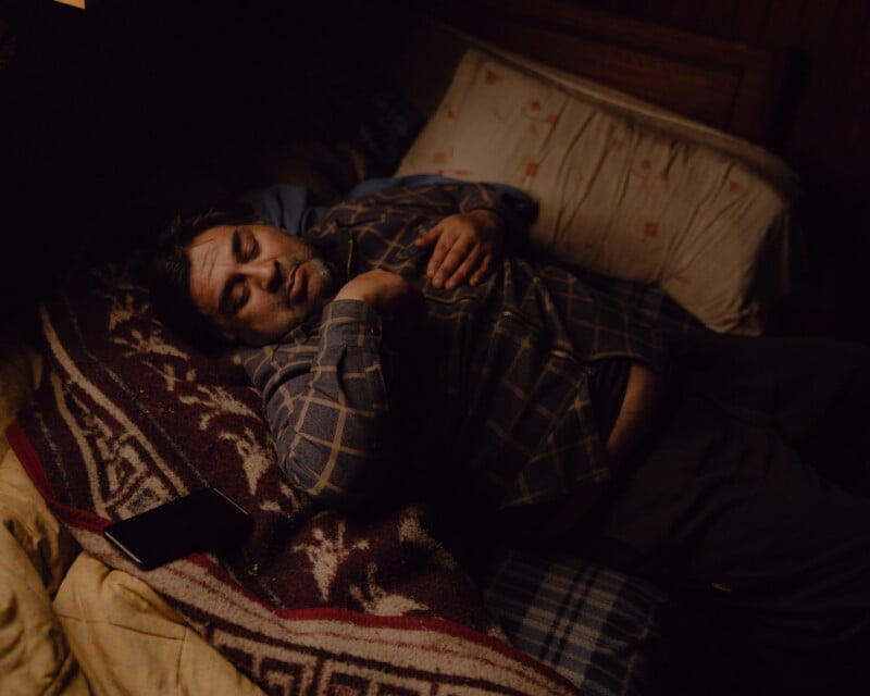 A man wearing a plaid shirt lies on a bed, resting on his back with one arm across his chest. A smartphone is next to him on a patterned blanket. The lighting is dim and warm.