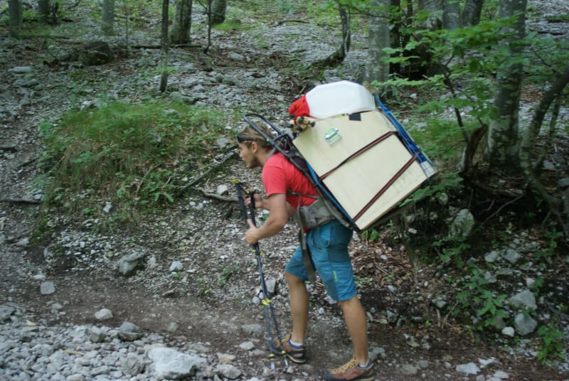 A person hiking on a rocky forest trail carries a large, bulky load on their back, including a cooler and various items, using hiking poles for support.