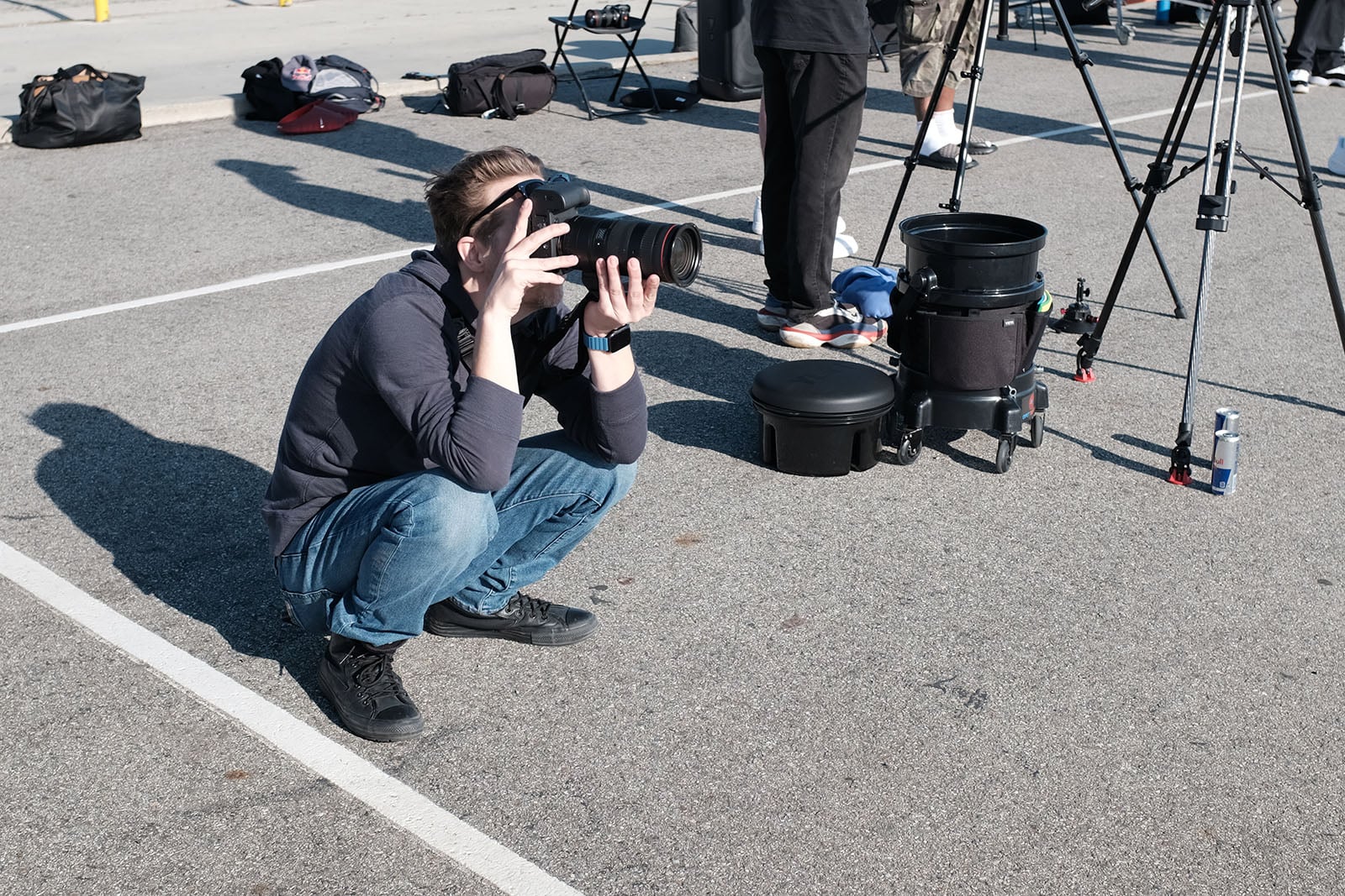 A person squats in a parking lot, holding a large camera with a telephoto lens. Tripods, camera equipment, and other people’s legs are visible in the background on the pavement.
