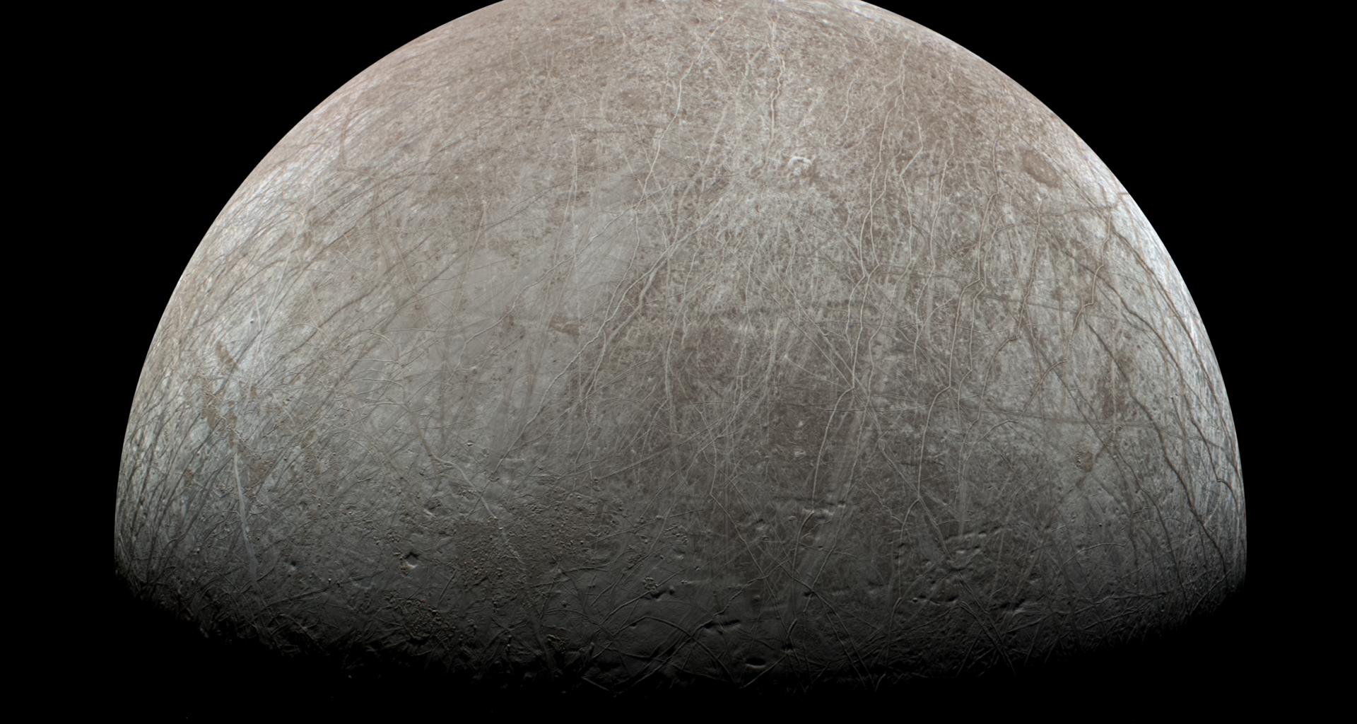 An image of the surface of Europa. The top half of the sphere is illuminated with the bottom half dark. The surface is traced with lineae, long lines across its surface of various hues of grey, white, and brown. The surface is a brown-grey, somewhat like Earth's Moon with the highest brightness areas appearing white.