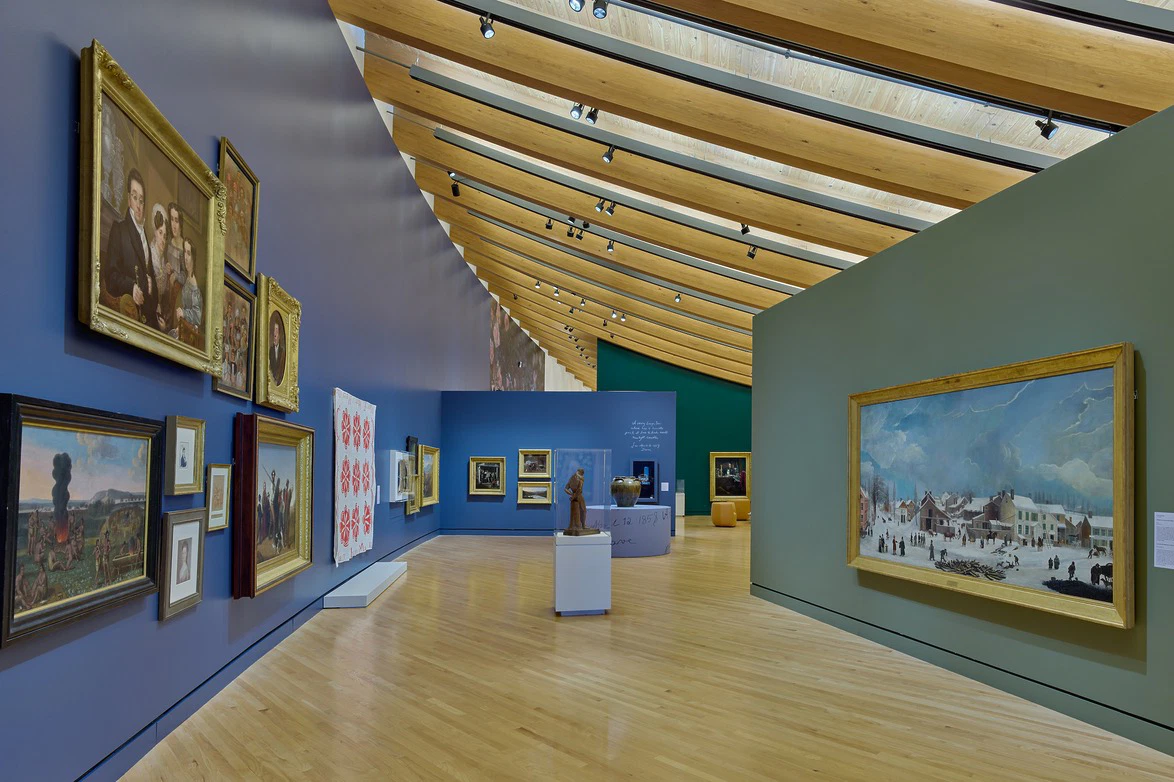 Art gallery with paintings on curved blue and green walls, wooden floor and ceiling, featuring various artworks and sculptures.
