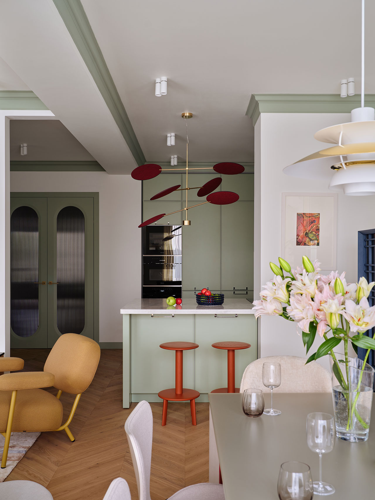Modern kitchen with mint green cabinets, red accents, and gold lighting fixture; dining area with flowers and wine glasses in the foreground.