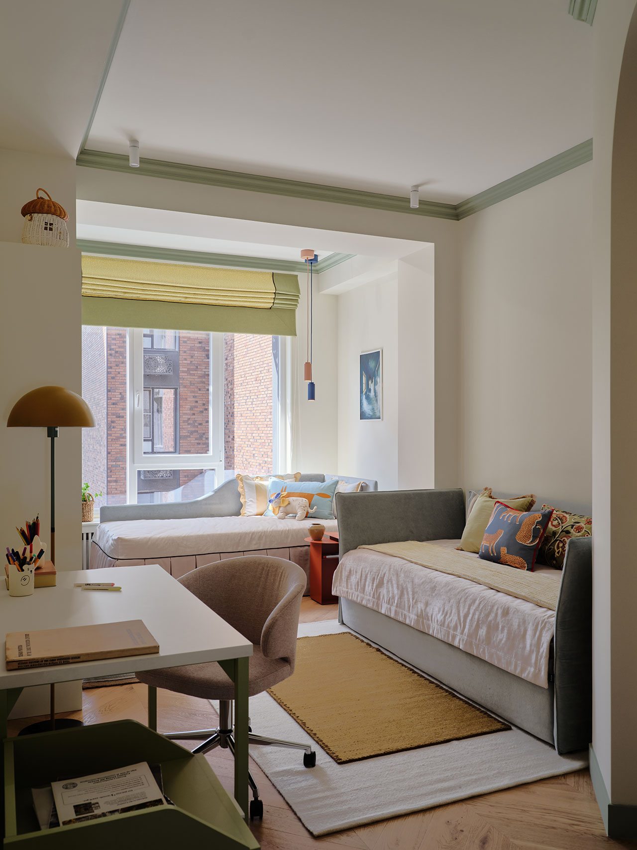 A modern, well-lit bedroom with two beds, a desk with a chair, a window with a city view, neutral walls, pastel decor, and a yellow rug on wooden flooring.