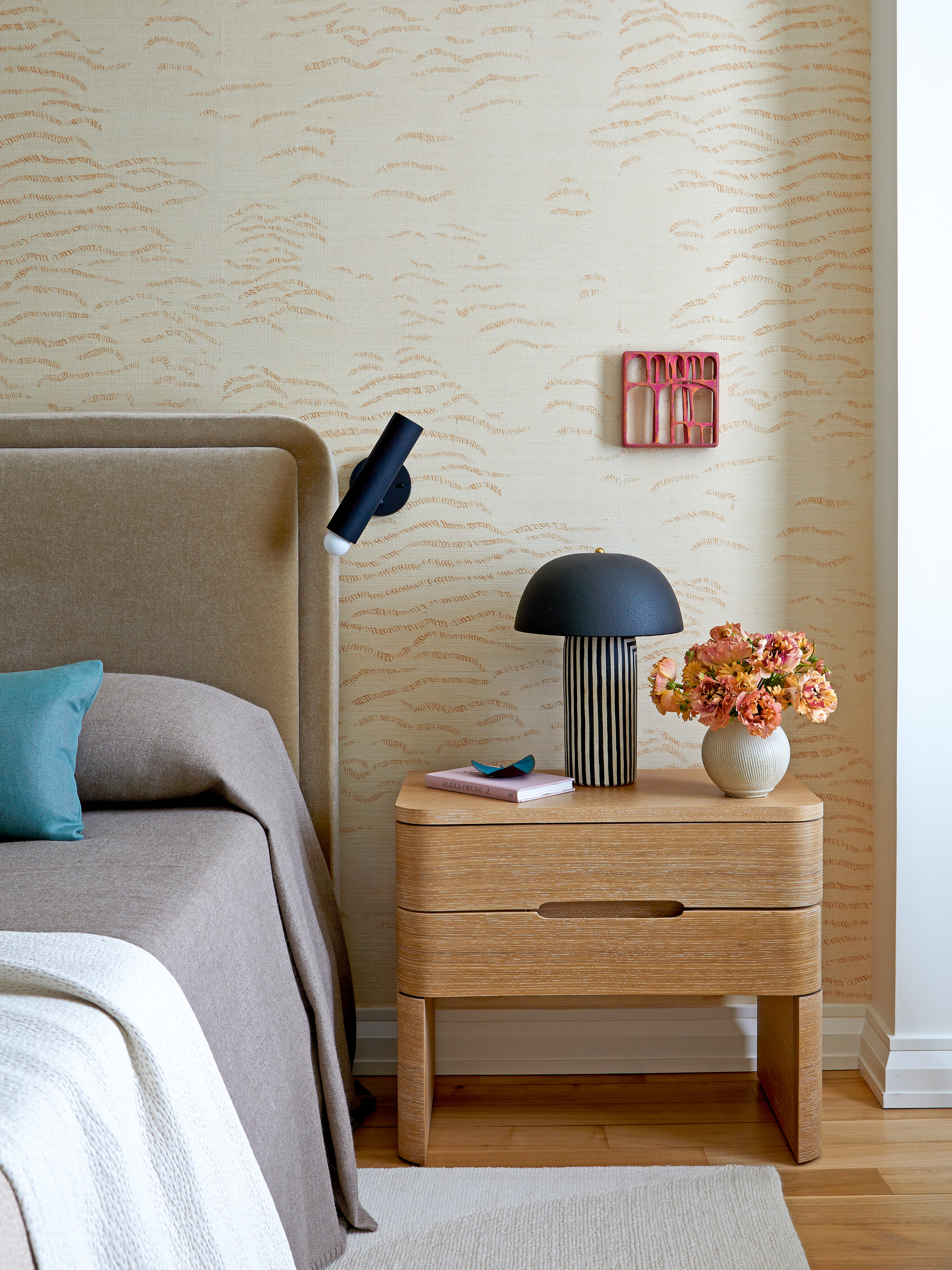 Bedroom with patterned beige wallpaper, greige upholstered headboard, wood side table and black and white striped lamp