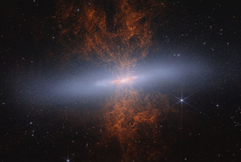 A spiral galaxy seen edge-on with glowing orange-red gas clouds extending above and below it; bright stars and distant galaxies are visible against the dark space background.