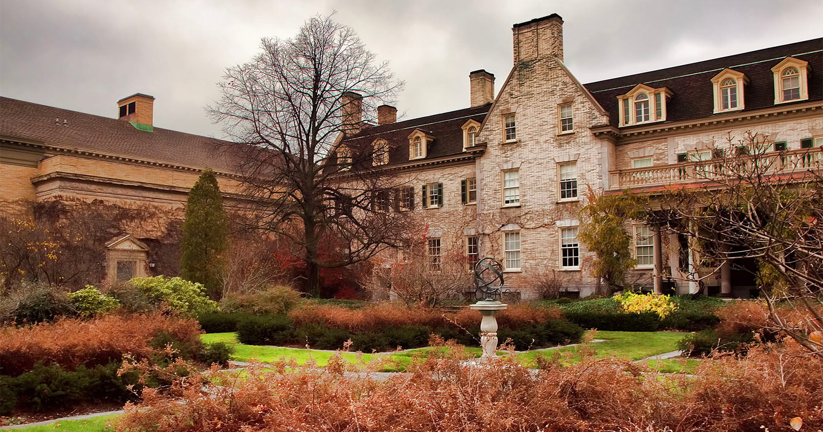 A large historic stone building with tall windows and dormers stands behind landscaped gardens with brown shrubs, winding paths, and a sundial under cloudy skies.