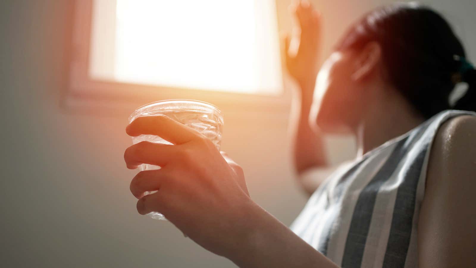 A woman holds a glass of water near a sunny window.