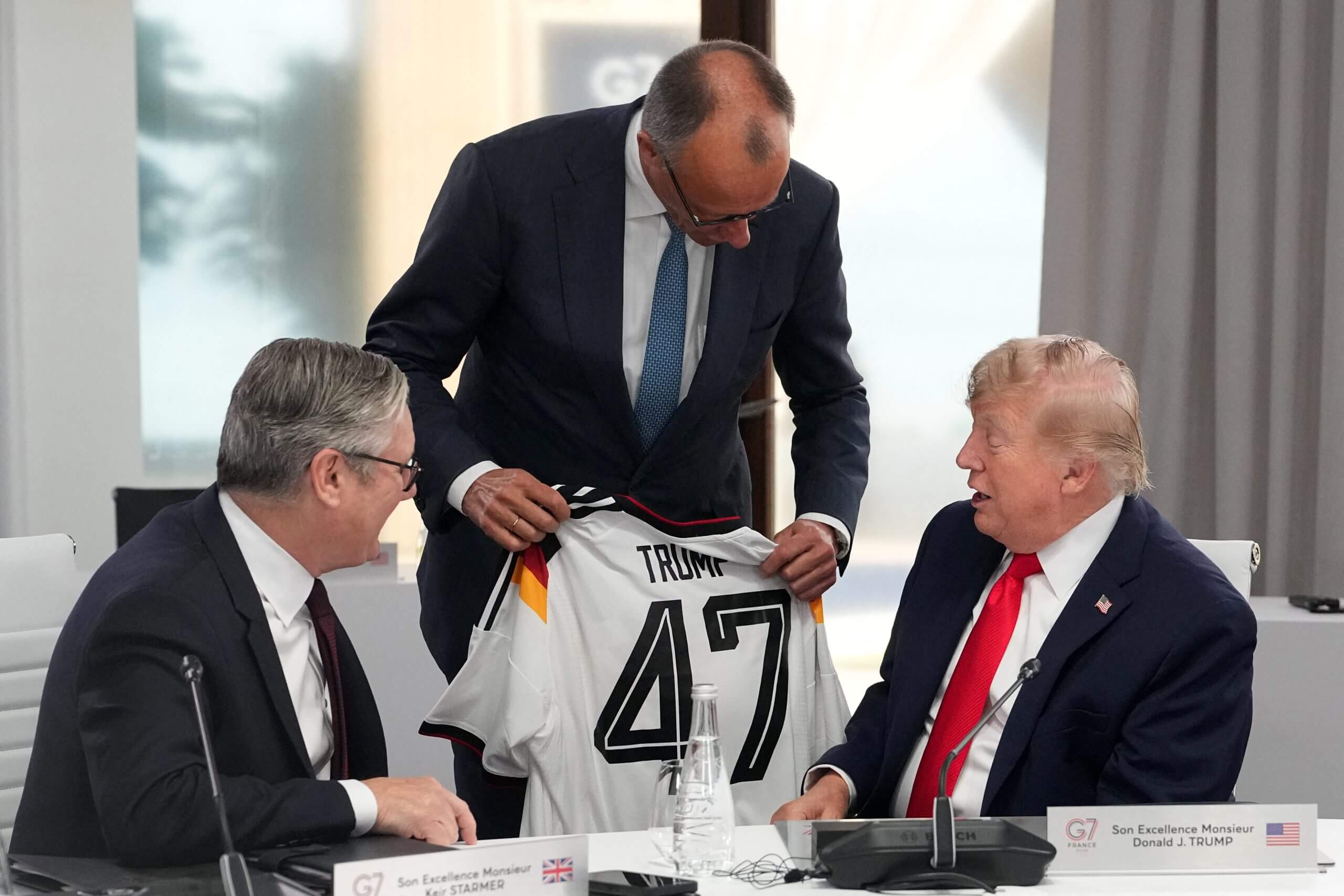 Two men sit either side of a man holding a white soccer jersey with '47' on the back