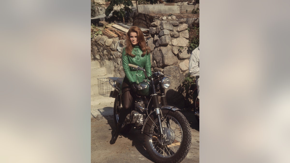 Ann-Margret sitting on a motorcycle wearing a green leather jacket and black tights outdoors