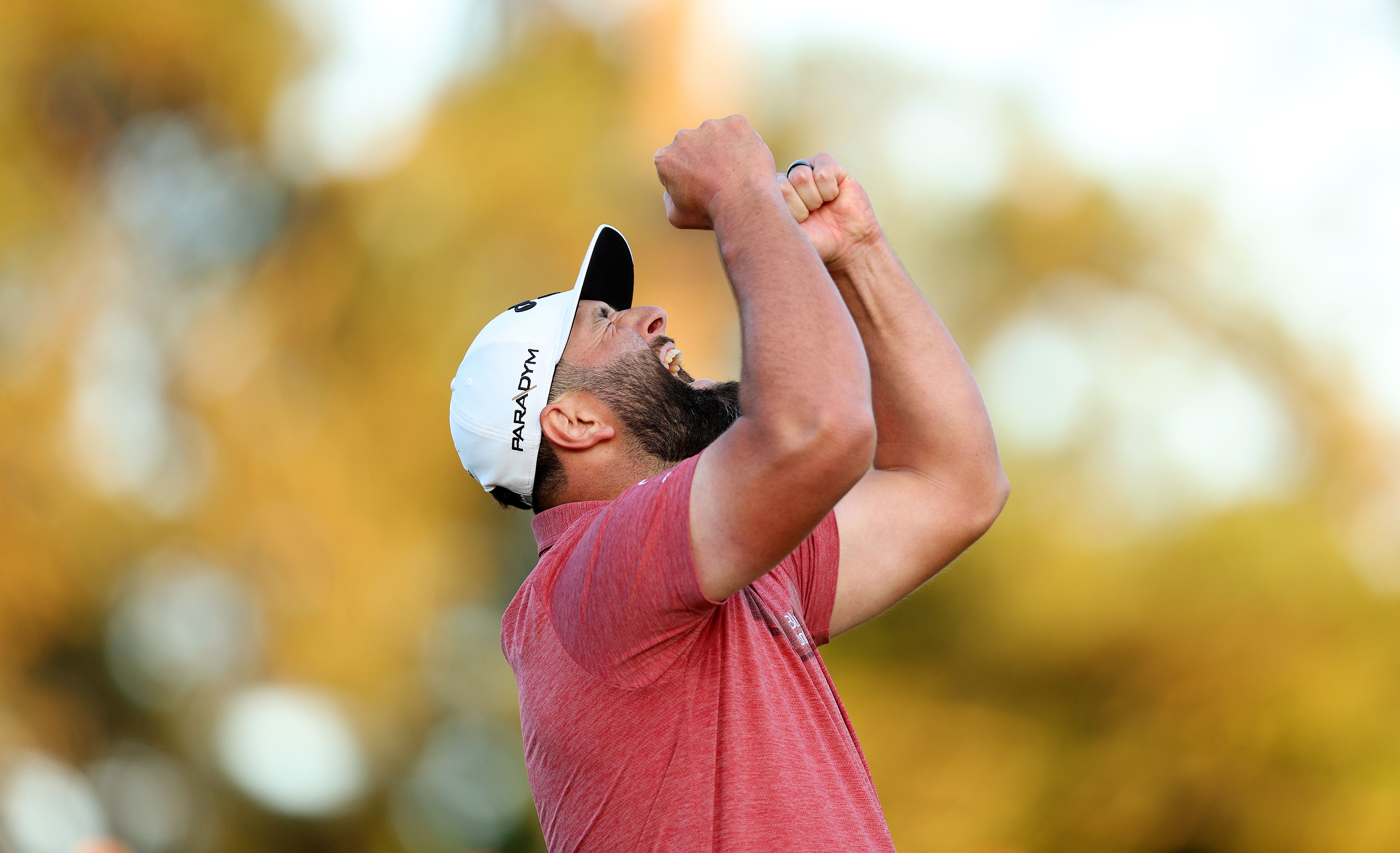 Golfer in red shirt and white cap celebrates with arms raised against a blurred background.