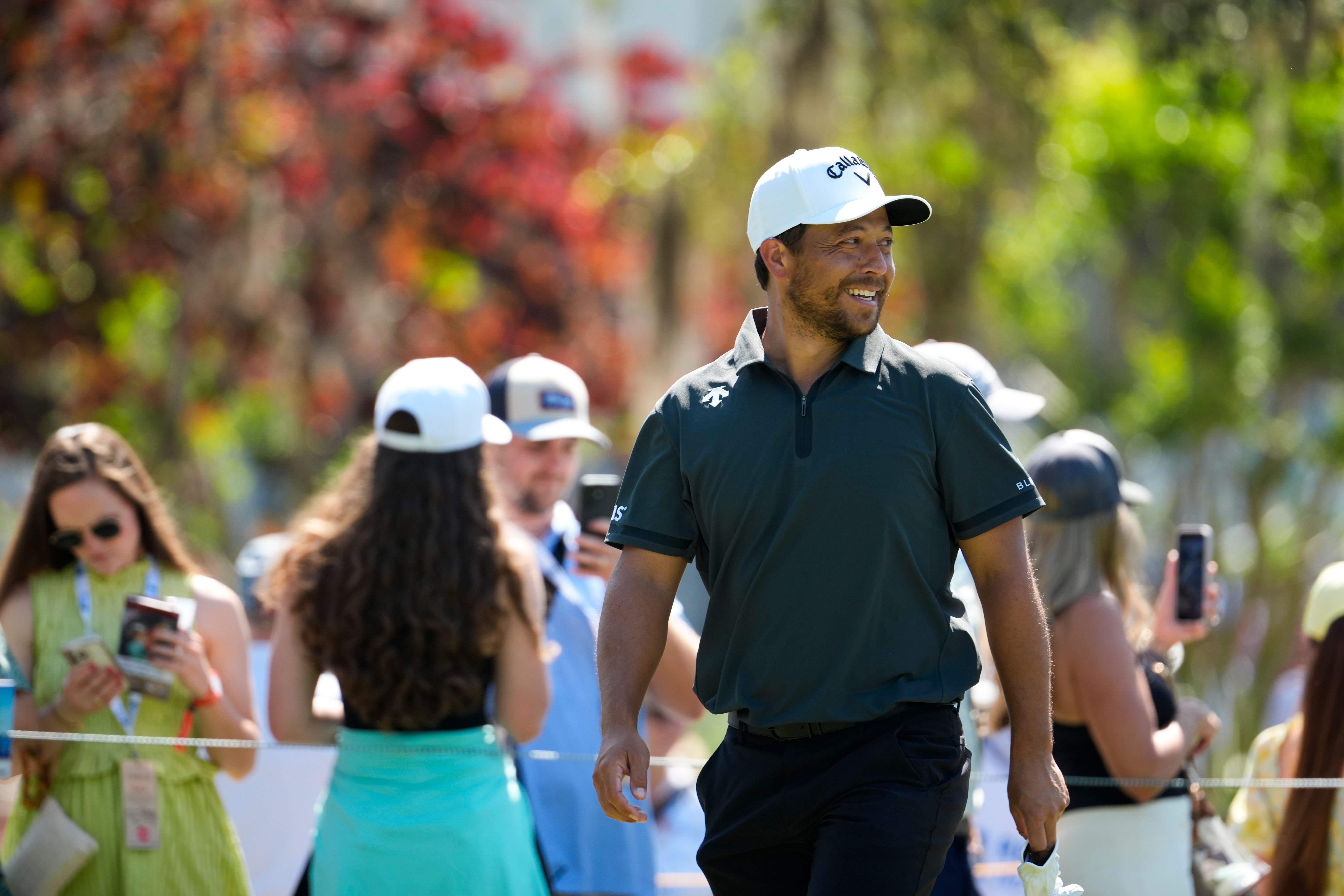 Golfer Tony Finau walks on a sunny course, smiling among spectators.